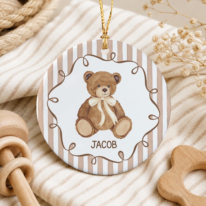 A round ceramic ornament featuring a brown teddy bear illustration with a cream-colored ribbon around its neck.