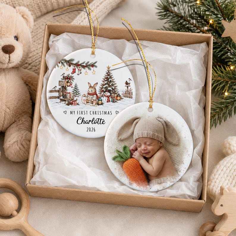 Two round white ceramic Christmas ornaments with gold hanging strings. One features a watercolor illustration of a rabbit, Christmas tree, and the text "MY FIRST CHRISTMAS Charlotte 2026".