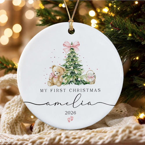 A round white ceramic ornament featuring a watercolor illustration of a teddy bear, a Christmas tree with a pink bow, and a basket. The text reads MY FIRST CHRISTMAS, amelia, and 2026, with two small pink baby footprints below.