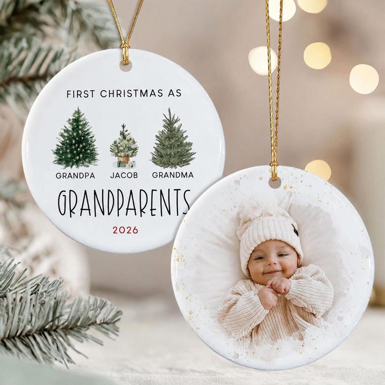 Two round white ceramic ornaments with gold hanging strings. One ornament features three watercolor pine trees with the text FIRST CHRISTMAS AS GRANDPA JACOB GRANDMA GRANDPARENTS 2026.
