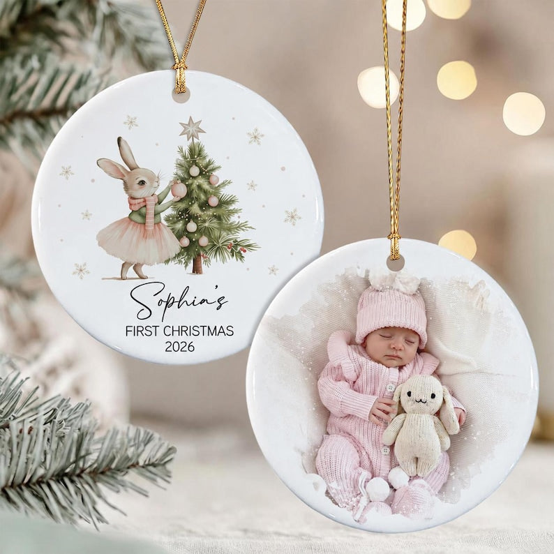 Two round white ceramic ornaments with gold hanging strings. One features a watercolor illustration of a rabbit in a pink tutu next to a Christmas tree with the text Sophia's FIRST CHRISTMAS 2026.