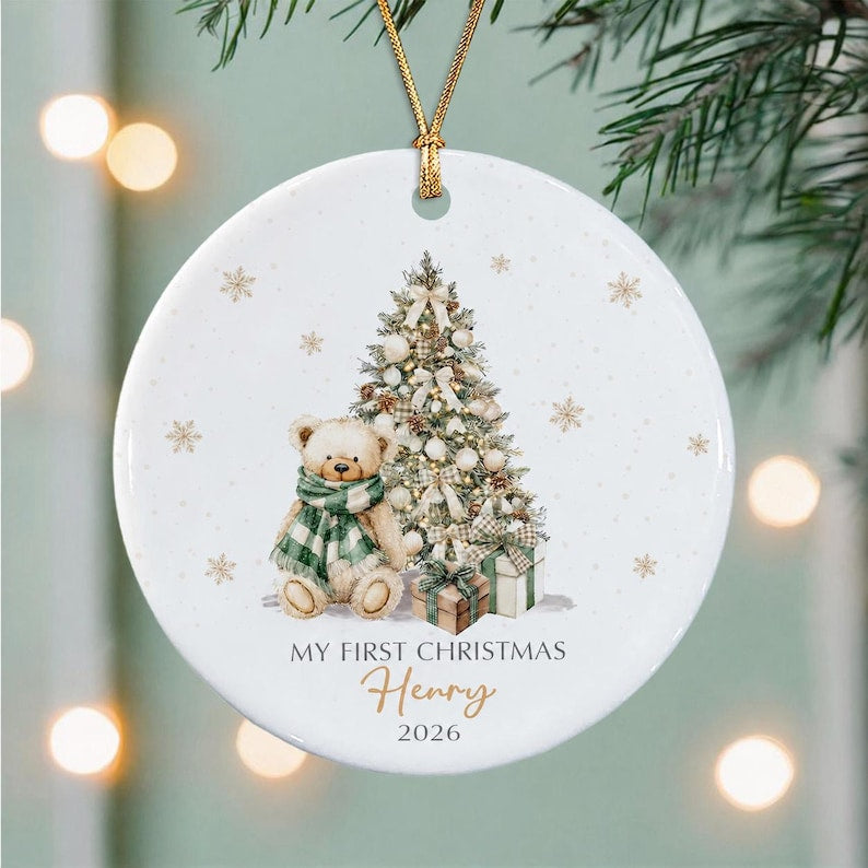 A round white ceramic Christmas ornament featuring a teddy bear wearing a green and white plaid scarf, a decorated Christmas tree, and wrapped gifts. The text reads MY FIRST CHRISTMAS Henry 2026 in black and gold lettering.