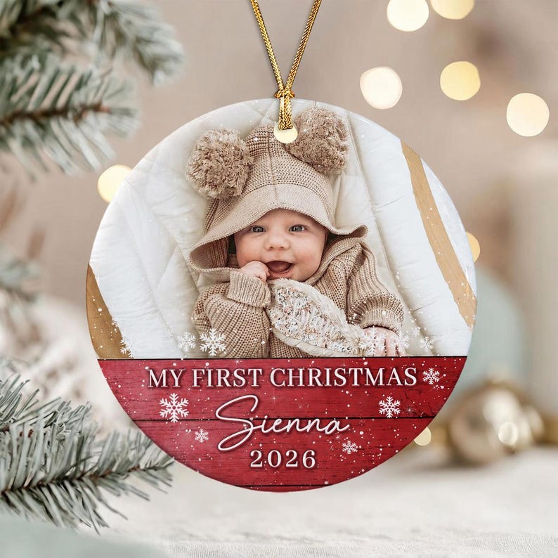 A round ceramic Christmas ornament featuring a photograph of a baby wearing a tan hooded sweater with pom-poms. The bottom section has a red wood-grain background with white snowflakes and the text My First Christmas Sienna 2026.