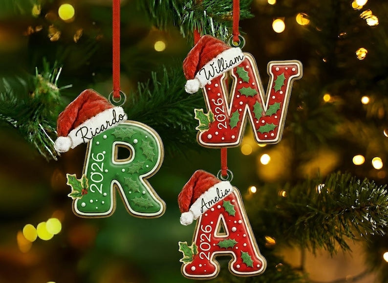 Three hanging Christmas ornaments shaped like the letters R, W, and A. Each letter features a red Santa hat, white icing dots, green holly leaves, and the year 2026. The letter R is green, while W and A are red.