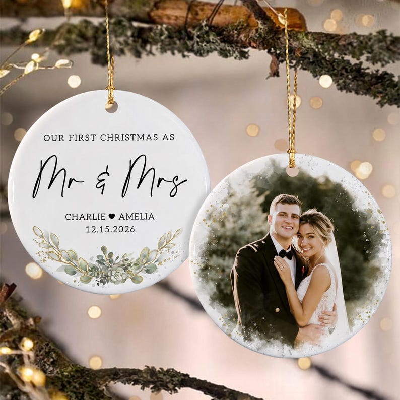 Two round white ceramic ornaments hanging by gold strings. One ornament features the black text "OUR FIRST CHRISTMAS AS Mr & Mrs", the names "CHARLIE & AMELIA", the date "12.15.2026", and a green watercolor botanical illustration.