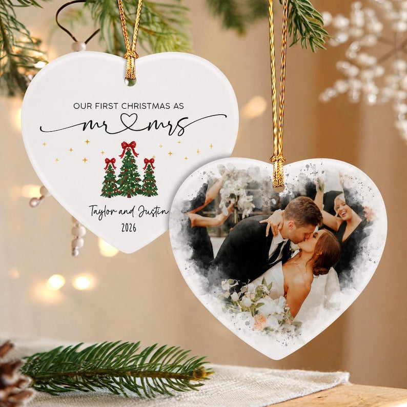 Two heart-shaped ceramic ornaments with gold hanging strings. One ornament features the text "OUR FIRST CHRISTMAS AS mr & mrs", "Taylor and Justin", "2026", and three Christmas trees.
