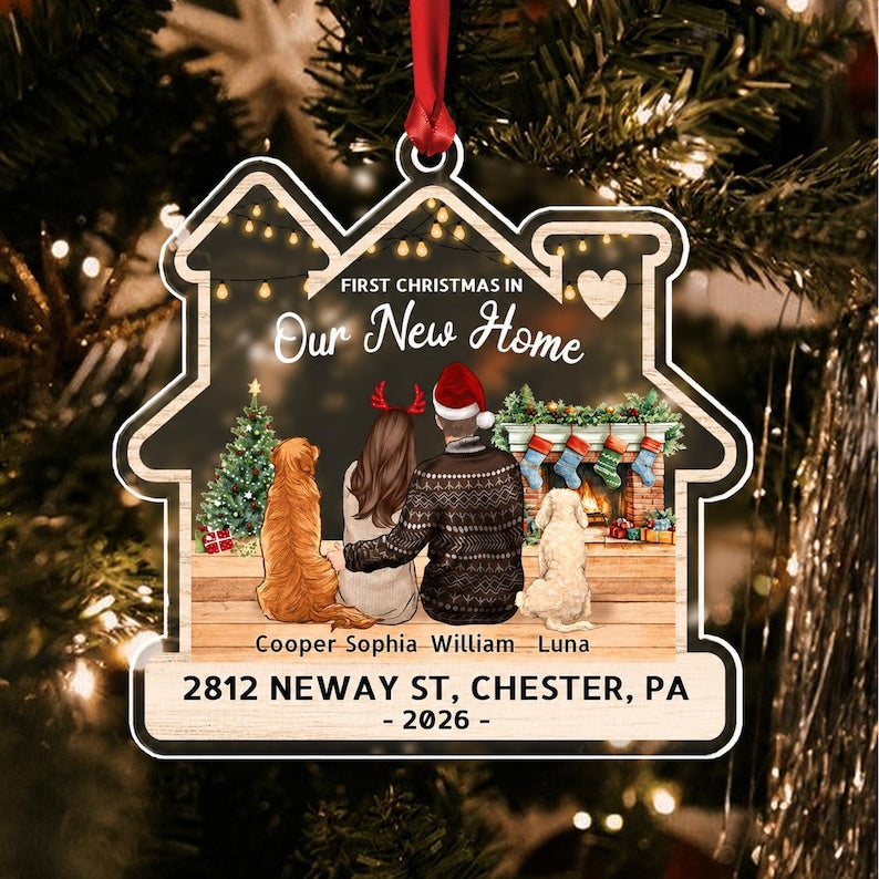 A house-shaped Christmas ornament featuring an illustration of two people and two dogs sitting by a fireplace. Text reads: FIRST CHRISTMAS IN Our New Home, Cooper, Sophia, William, Luna, 2812 NEWAY ST, CHESTER, PA - 2026 -.