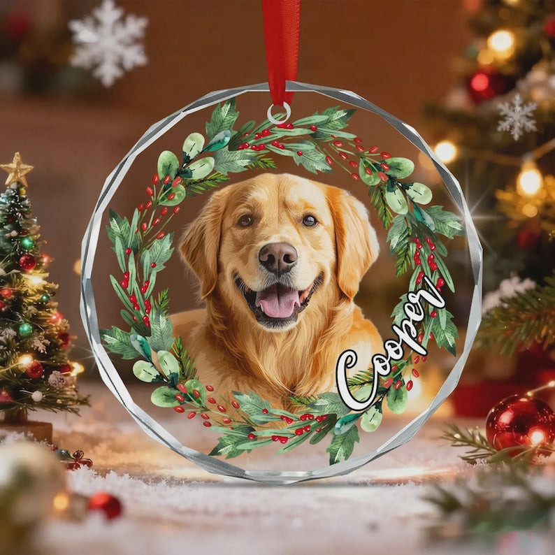 A circular clear acrylic Christmas ornament featuring a golden retriever portrait framed by a watercolor wreath of green leaves and red berries. The name Cooper is written in a white cursive script on the right side of the wreath.