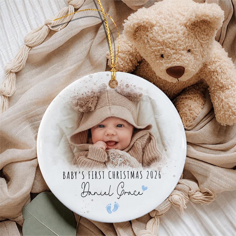 A round white ceramic ornament featuring a photograph of a baby wearing a tan hooded sweater. The text reads "BABY'S FIRST CHRISTMAS 2026" and "Daniel Grace" in black script, with two small blue footprints and a blue heart icon.