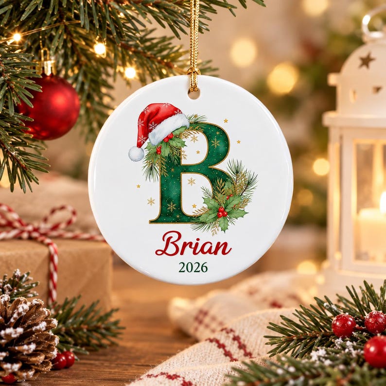 A round white ceramic ornament featuring a large green letter B decorated with pine needles, holly berries, and a red Santa hat. The name Brian is printed in red script below the letter, followed by the year 2026 in black text.