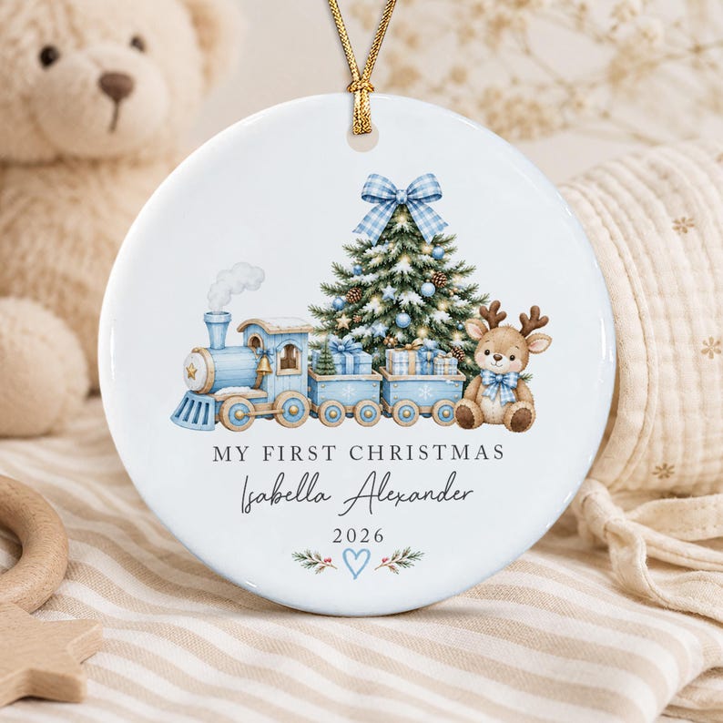 A round white ceramic ornament featuring a blue train, a decorated Christmas tree, and a reindeer. The text reads MY FIRST CHRISTMAS, Isabella Alexander, and 2026, with a blue heart and pine branches at the bottom.