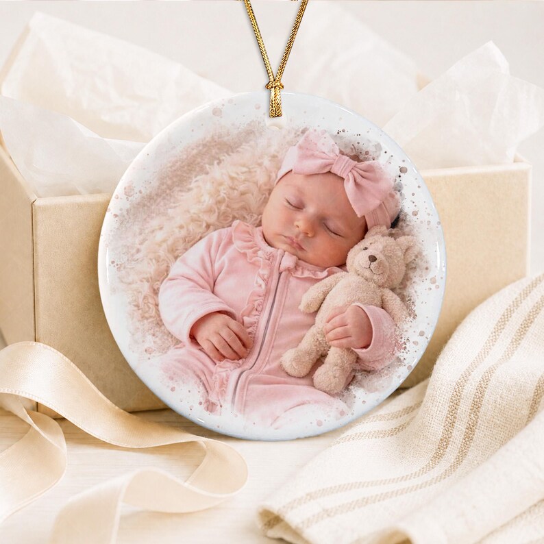 A round white ceramic ornament featuring a printed photograph of a sleeping infant wearing a light pink outfit and a matching bow headband, holding a small tan teddy bear. The ornament hangs from a thin gold cord.
