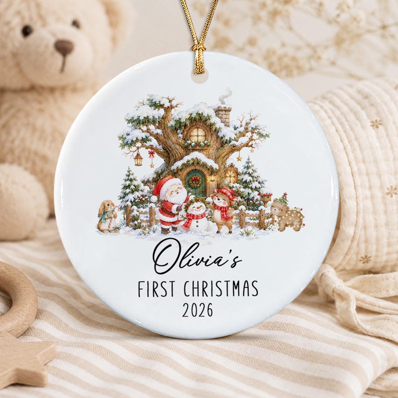 A round white ceramic ornament featuring a festive illustration of a snow-covered tree house, Santa Claus, a snowman, and woodland animals. The text reads Olivia's FIRST CHRISTMAS 2026.