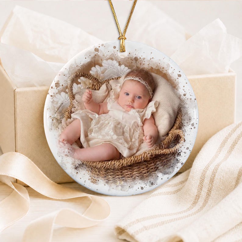 A round white ceramic ornament featuring a printed photograph of an infant wearing a white lace dress and headband, resting in a woven basket. The ornament is suspended by a thin gold cord and has a speckled brown border design.