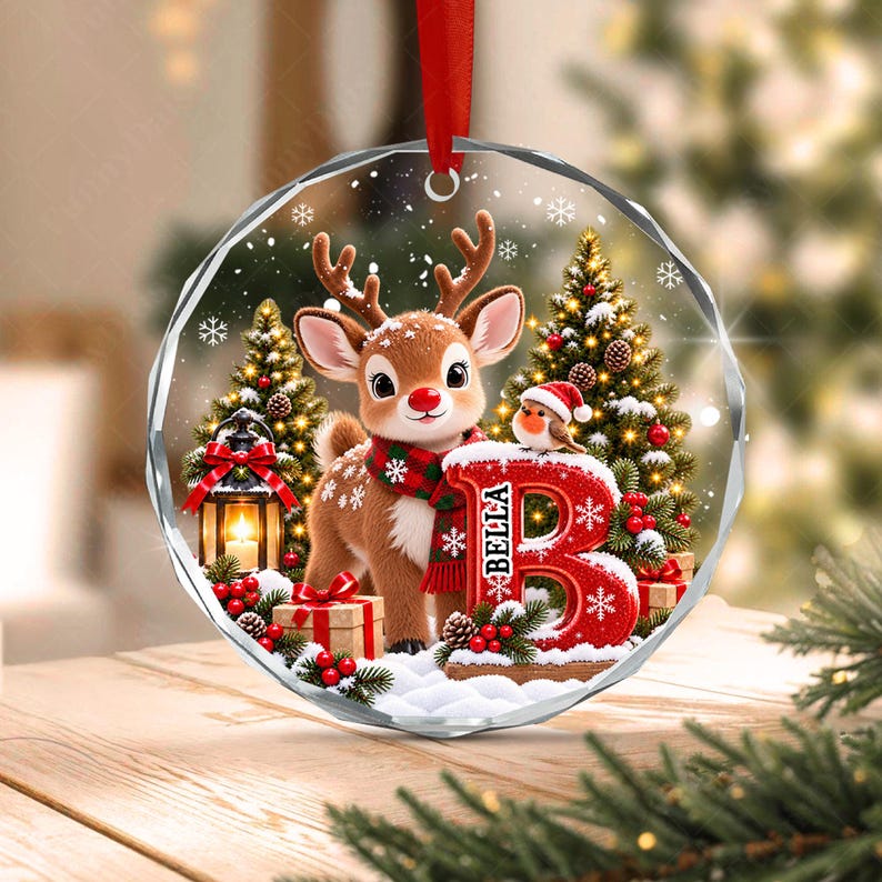 A round, clear acrylic Christmas ornament featuring a printed illustration of a brown reindeer wearing a red and green plaid scarf, standing next to a large red letter B with the name Bella, a small bird, and a lantern.