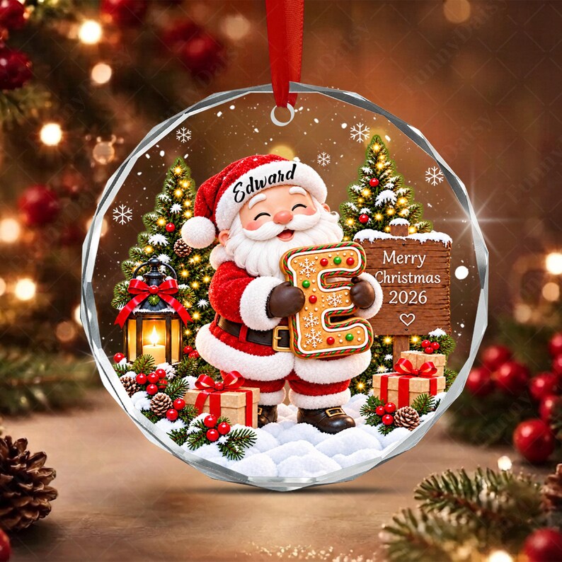 A round, faceted glass Christmas ornament featuring a cartoon Santa Claus holding a large gingerbread letter E.