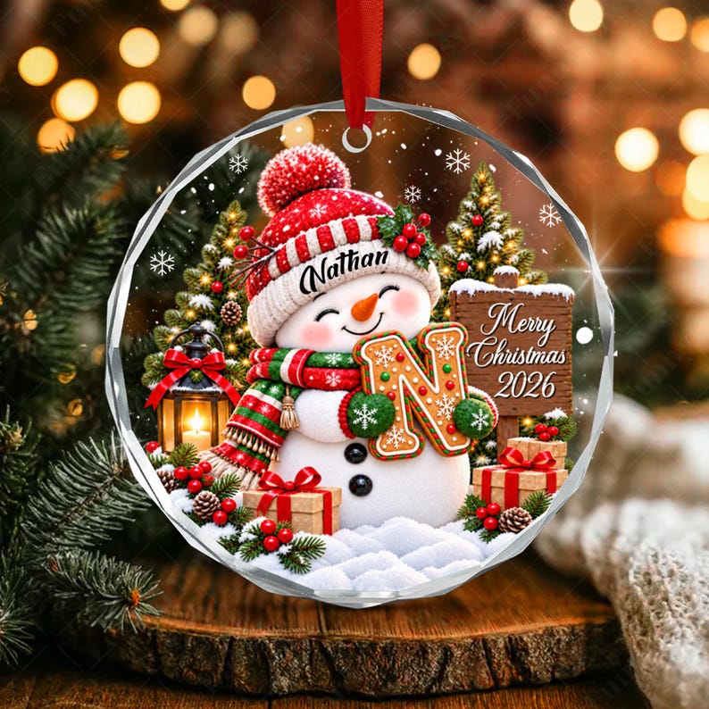 A round, clear acrylic Christmas ornament featuring a snowman wearing a red and white knit hat with the name Nathan, a green and red scarf, and holding a gingerbread letter N. A wooden sign reads Merry Christmas 2026.