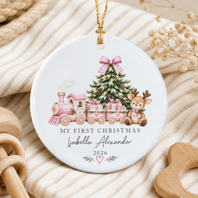 A round white ceramic ornament featuring a pink train, a Christmas tree, and a reindeer. The text reads MY FIRST CHRISTMAS, Isabella Alexander, and 2026, with a small heart and pine branch graphic at the bottom.