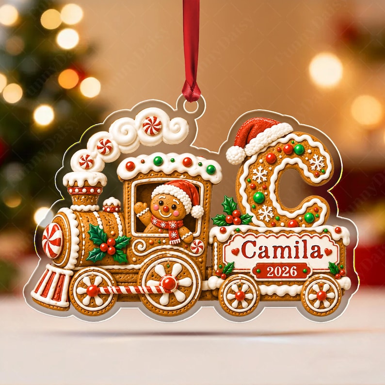 A gingerbread train Christmas ornament hanging from a red ribbon. The train features a gingerbread person in the cabin, a large letter C with a Santa hat, and a carriage labeled with the name Camila and the year 2026.