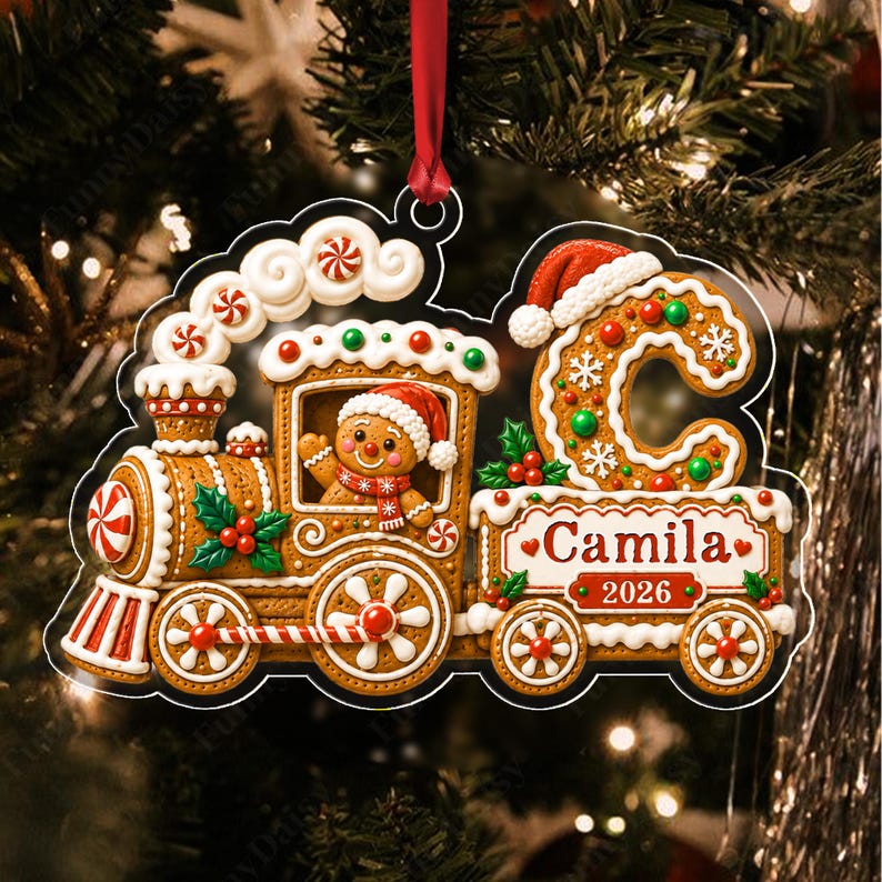 A gingerbread train ornament featuring a gingerbread person in a Santa hat, a large letter C with white icing snowflakes, and a red ribbon hanger. The train displays the name Camila and the year 2026 in red text on a white panel.