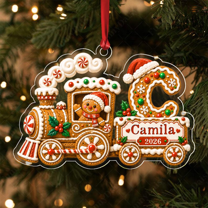 A gingerbread train ornament with a red ribbon, featuring a gingerbread person in a Santa hat, a large letter C, and the name Camila with the year 2026 printed on the side. The design includes white icing, red and green accents.