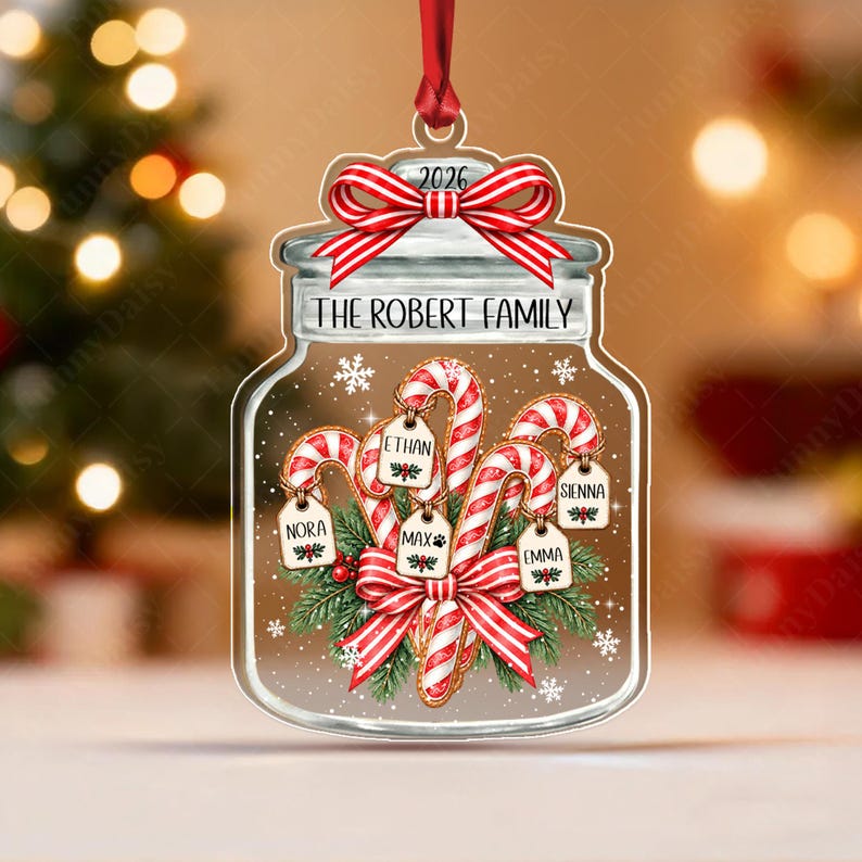 A clear jar-shaped Christmas ornament featuring a red ribbon bow at the top with the year 2026. The jar is labeled The Robert Family and contains candy canes with small wooden tags labeled Ethan, Sienna, Nora, Max, and Emma.