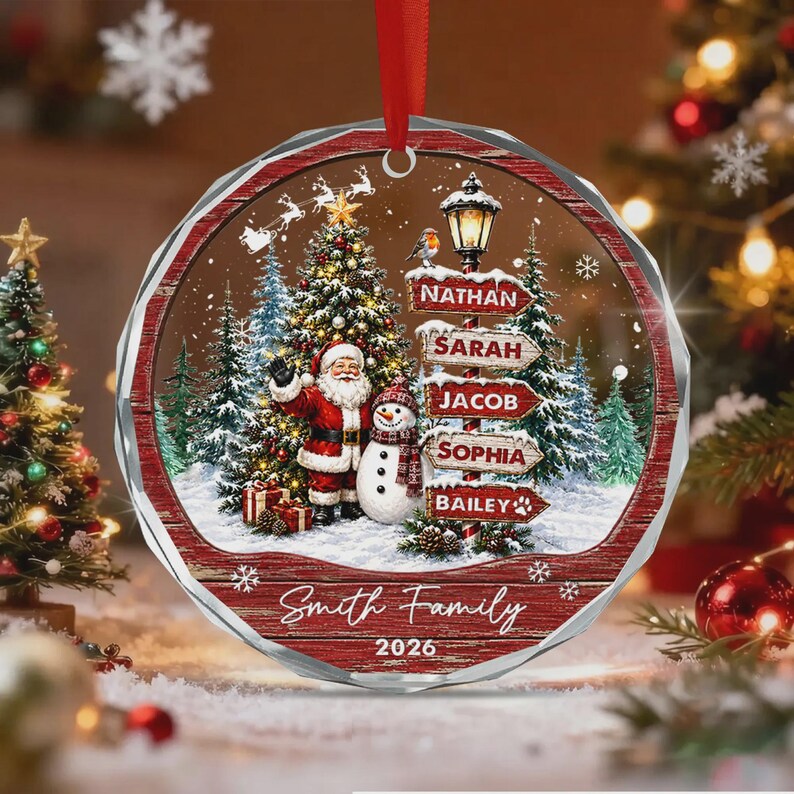 A round, clear acrylic Christmas ornament featuring a festive winter scene with Santa Claus, a snowman, and a street sign listing the names Nathan, Sarah, Jacob, Sophia, and Bailey. The text Smith Family and 2026 is printed below.