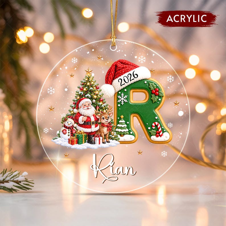 A round clear acrylic ornament featuring a green letter R wearing a red Santa hat with the year 2026 printed on it. The design includes a Christmas tree, Santa Claus, a reindeer, a snowman, gifts, and the name Rian in script text.