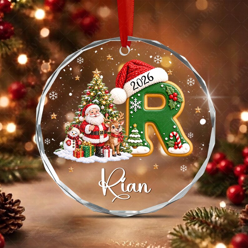 A circular glass Christmas ornament featuring a green letter R wearing a red Santa hat with the year 2026, a decorated Christmas tree, Santa Claus, a reindeer, a snowman, and the name Rian printed in white script on the bottom.