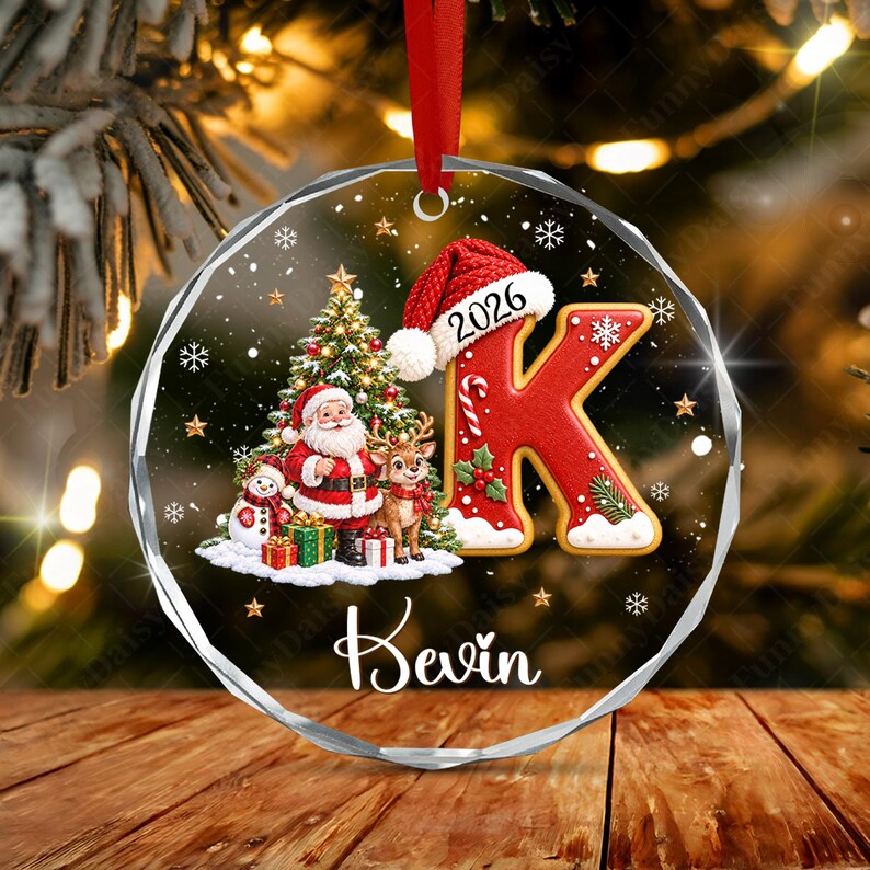 A round, clear acrylic Christmas ornament featuring a red letter K wearing a Santa hat with the year 2026, a Santa Claus figure, a reindeer, a snowman, gifts, and a Christmas tree. The name Kevin is printed in white script below.