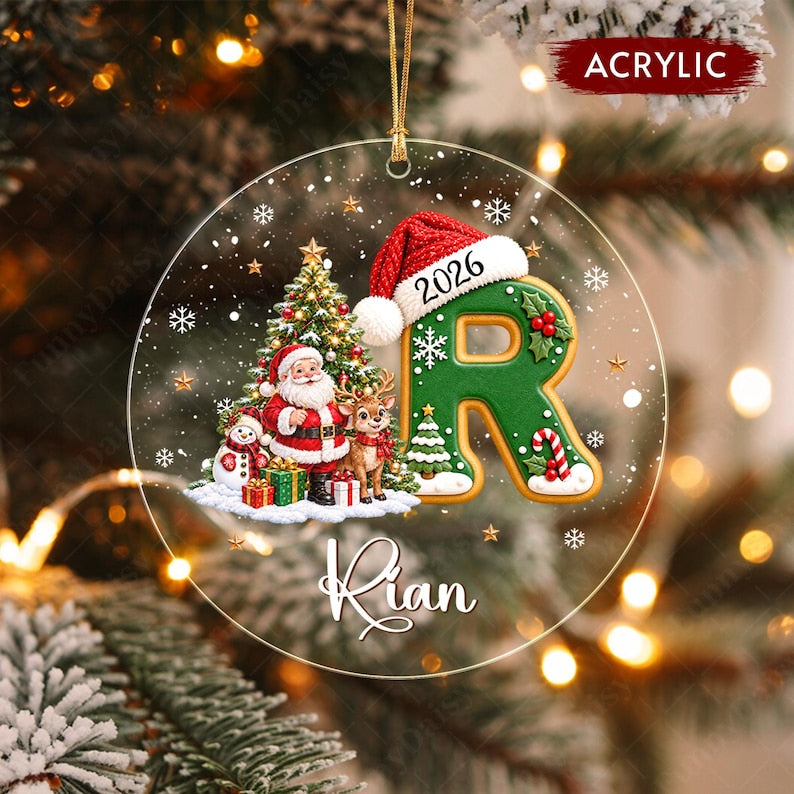 A round clear acrylic Christmas ornament featuring a green letter R wearing a Santa hat with the year 2026, a Santa Claus figure, a reindeer, a snowman, a Christmas tree, and the name Rian printed in white script lettering.