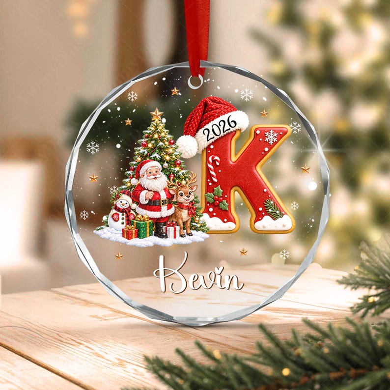 A circular clear acrylic ornament featuring a red letter K wearing a Santa hat with the year 2026, a Christmas tree, Santa Claus, a reindeer, a snowman, and gifts.