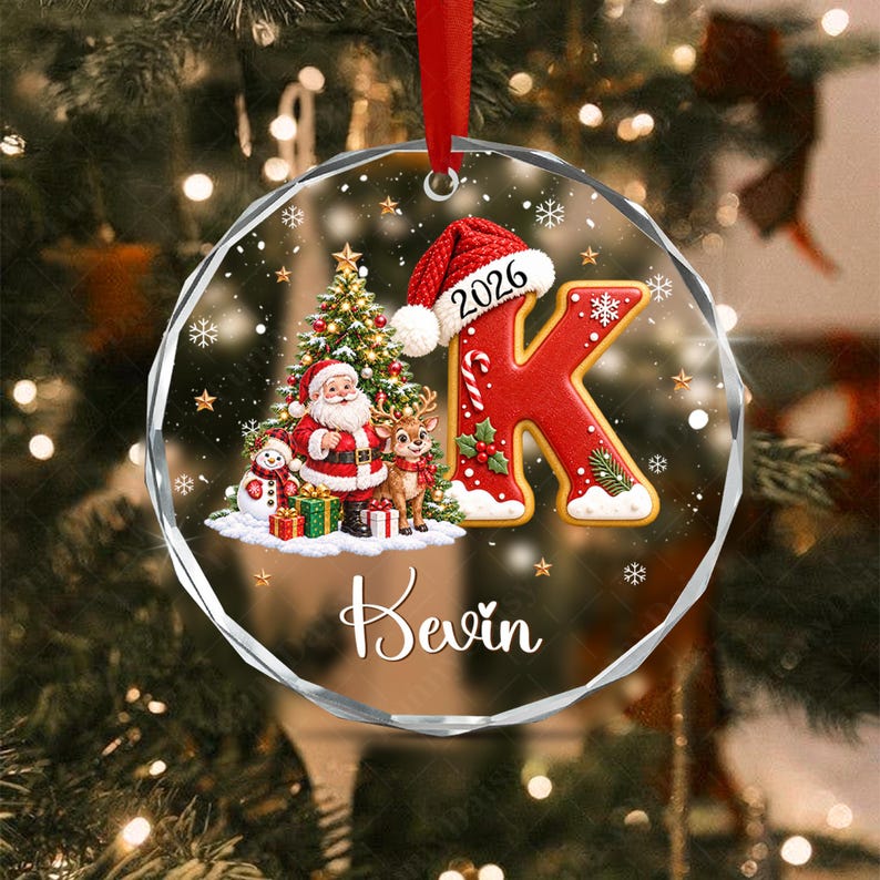 A round, faceted glass Christmas ornament featuring a red letter K wearing a Santa hat with the year 2026, a decorated Christmas tree, Santa Claus, a reindeer, a snowman, and the name Kevin printed in white script on a red ribbon.