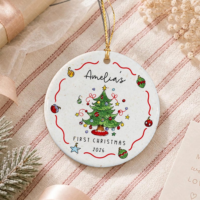 A round white ceramic Christmas ornament featuring a hand-drawn green Christmas tree in the center, surrounded by colorful ornaments and stars. The text reads Amelia's FIRST CHRISTMAS 2026. It hangs from a gold string.