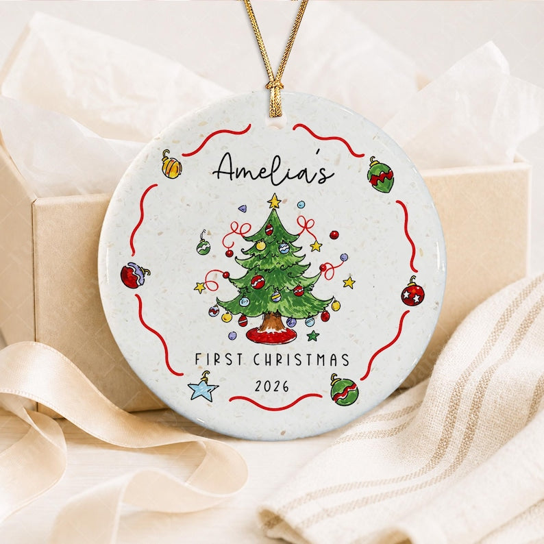 A round white ceramic ornament featuring a hand-drawn green Christmas tree with colorful baubles and stars. The text reads Amelia's FIRST CHRISTMAS 2026. The design is framed by a red wavy line and small decorative ornaments.