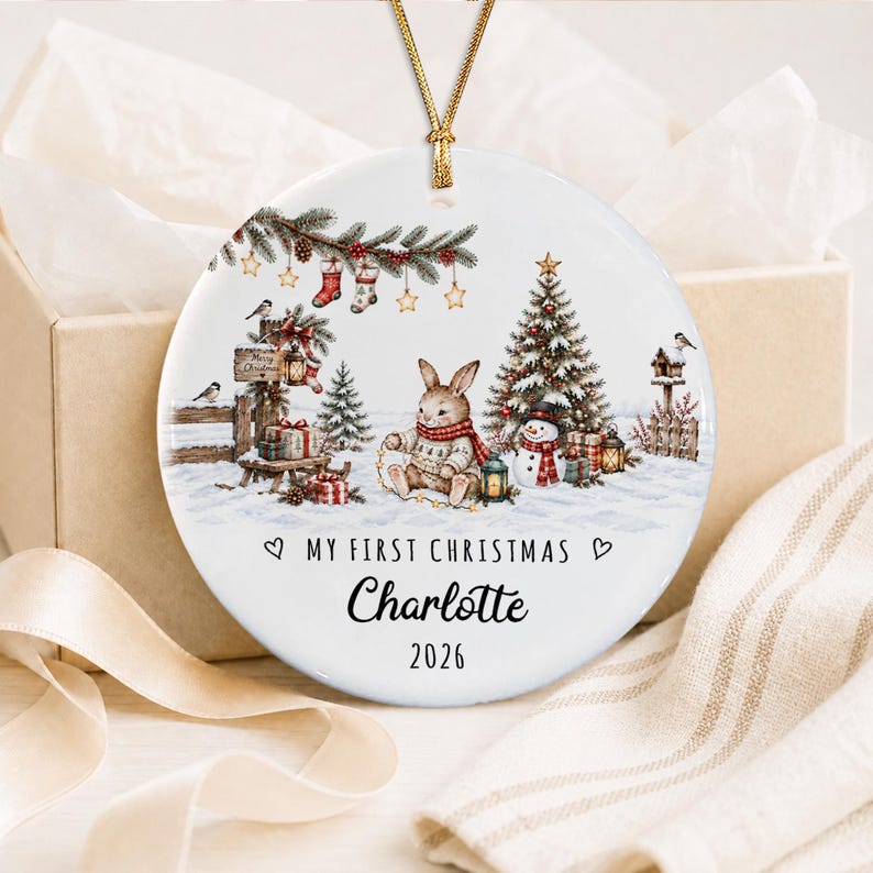 A round white ceramic ornament hanging from a gold cord, featuring a festive illustration of a rabbit, a Christmas tree, and a snowman in a snowy landscape. The text reads MY FIRST CHRISTMAS, Charlotte, and the year 2026.