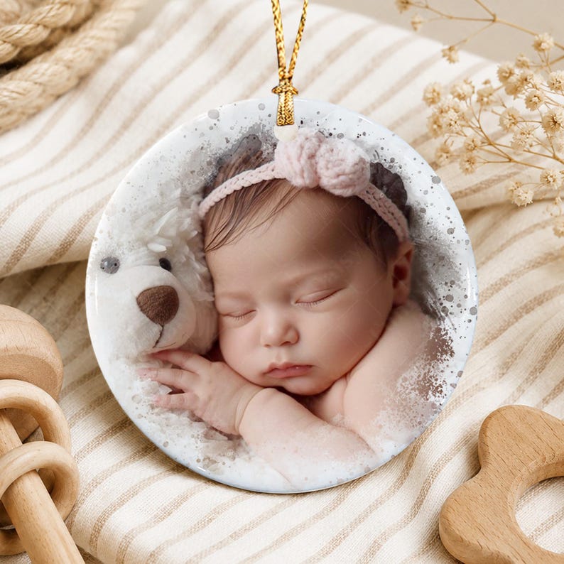 A round ceramic ornament featuring a printed photograph of a sleeping infant wearing a light pink headband with a bow, resting against a white plush teddy bear, finished with a gold hanging cord and a speckled gray border design.