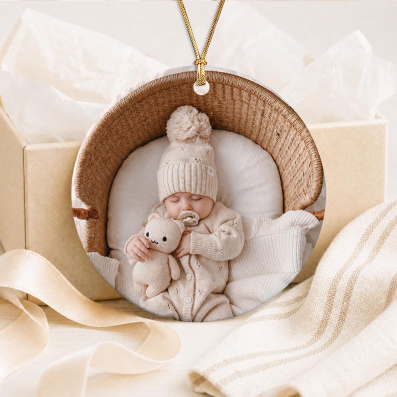 A circular ceramic ornament featuring a printed photograph of a baby wearing a beige knit hat and matching onesie, holding a small plush toy while resting in a woven basket. The ornament hangs from a gold-colored cord.