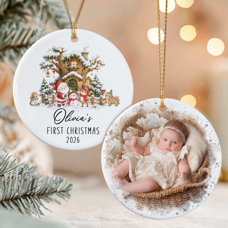 Two round white ceramic ornaments with gold hanging strings. One features a winter forest scene with a tree house, Santa, and animals, plus the text Olivia's FIRST CHRISTMAS 2026.