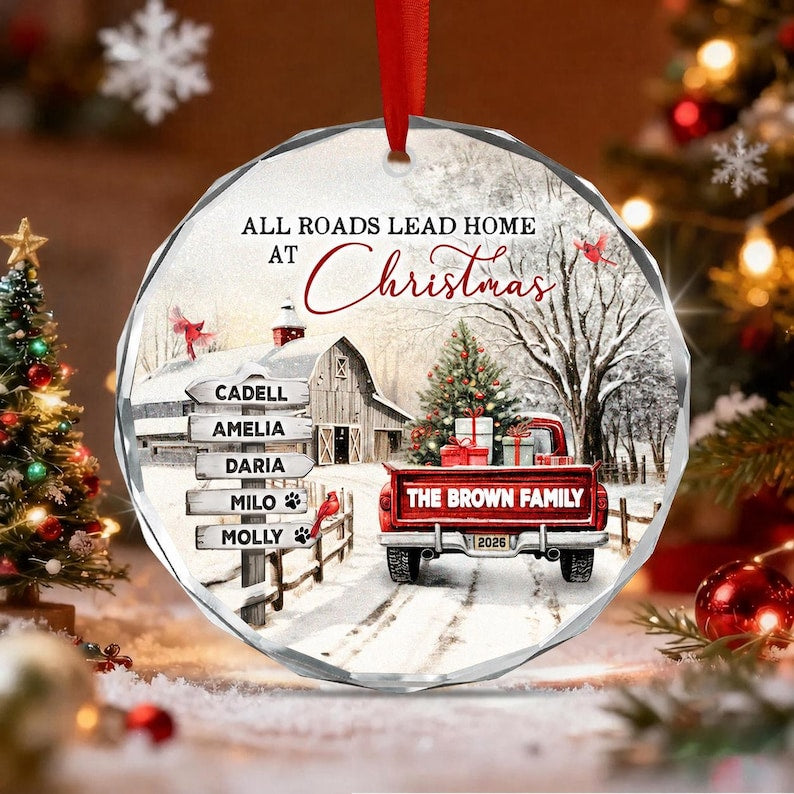 A round, faceted glass Christmas ornament featuring a winter scene with a red truck, a barn, and a signpost with the names Cadell, Amelia, Daria, Milo, and Molly.