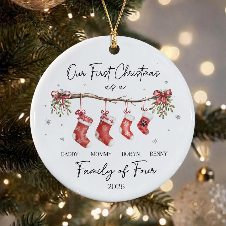 A round white ceramic Christmas ornament featuring the text Our First Christmas as a Family of Four 2026. The design includes a branch with four hanging red stockings, mistletoe, and the names Daddy, Mommy, Robyn, and Benny.