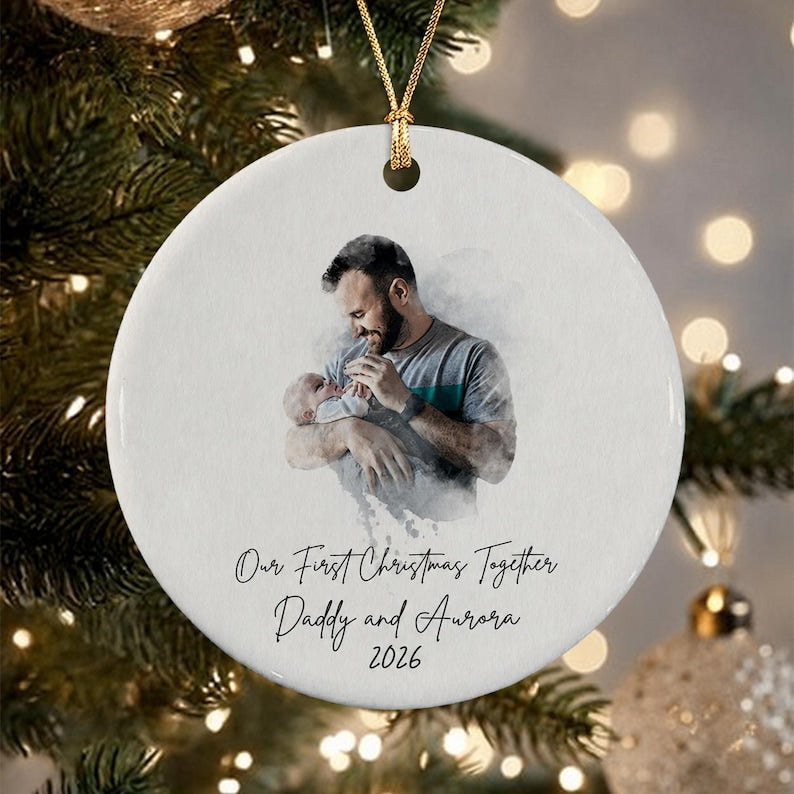 A round white ceramic ornament featuring a watercolor-style illustration of a parent holding a baby. Below the image, black cursive text reads: Our First Christmas Together, Daddy and Aurora, 2026.
