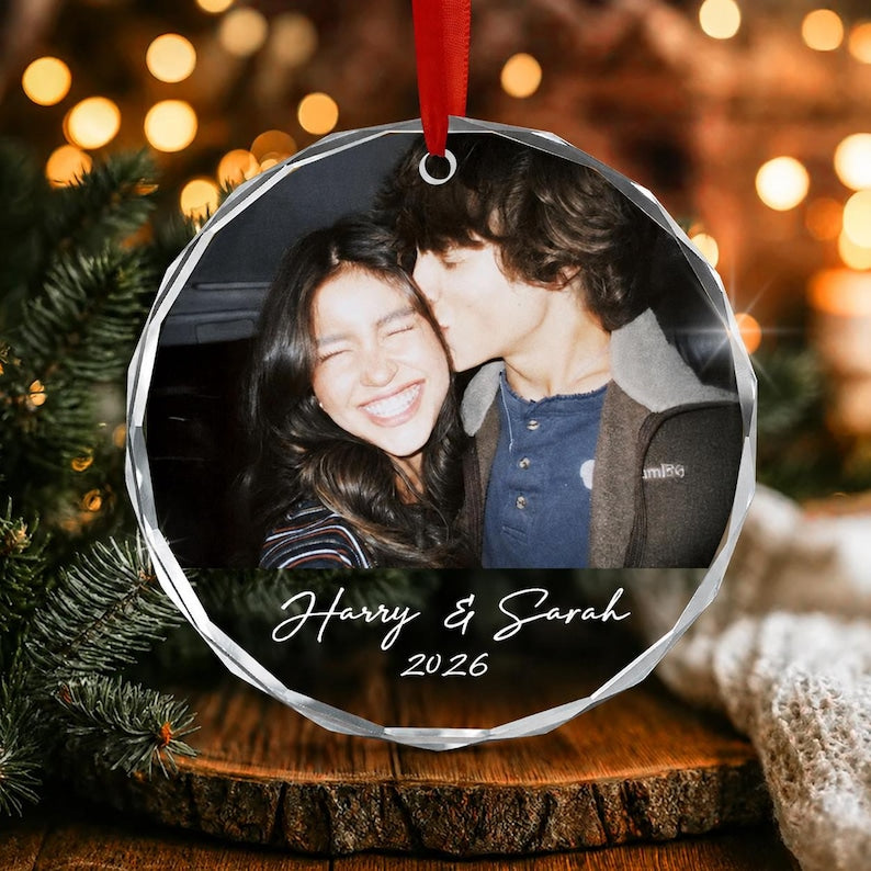 A round, faceted glass ornament featuring a printed photograph of two people, with the text Harry & Sarah and the year 2026 printed in white script below the image. The ornament hangs from a red ribbon loop at the top.
