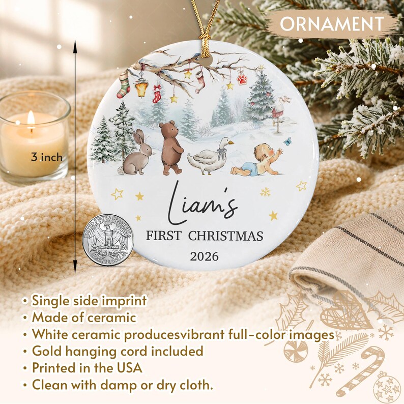 A round white ceramic ornament with a gold hanging cord, measuring 3 inches in diameter. The design features a winter forest scene with a rabbit, bear, goose, and baby, with the text 'Liam's FIRST CHRISTMAS 2026' printed below.