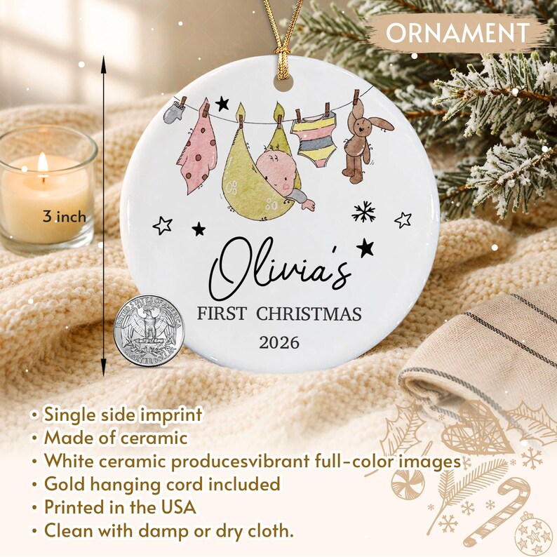 A round white ceramic ornament featuring a baby illustration on a clothesline with the text Olivia's FIRST CHRISTMAS 2026. A visible ruler indicates a 3 inch diameter next to a US quarter coin for scale with a gold hanging cord.