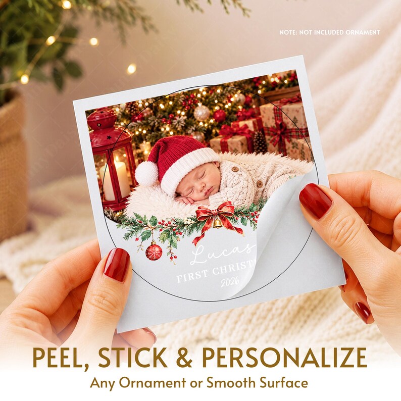 A square adhesive decal featuring a circular design with a photograph of a sleeping infant in a red Santa hat, surrounded by Christmas decorations, a red bow, and the text Lucas FIRST CHRISTMAS 2026, held by two hands.