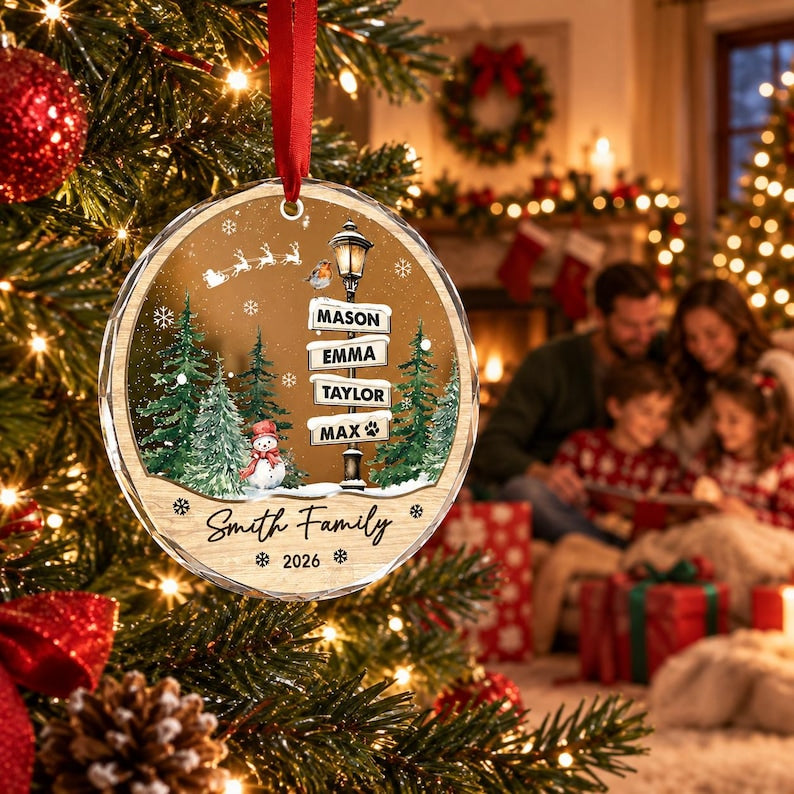 A round, clear acrylic Christmas ornament hanging from a red ribbon. The design features a winter scene with a street lamp, a snowman, pine trees, and a silhouette of Santa's sleigh.