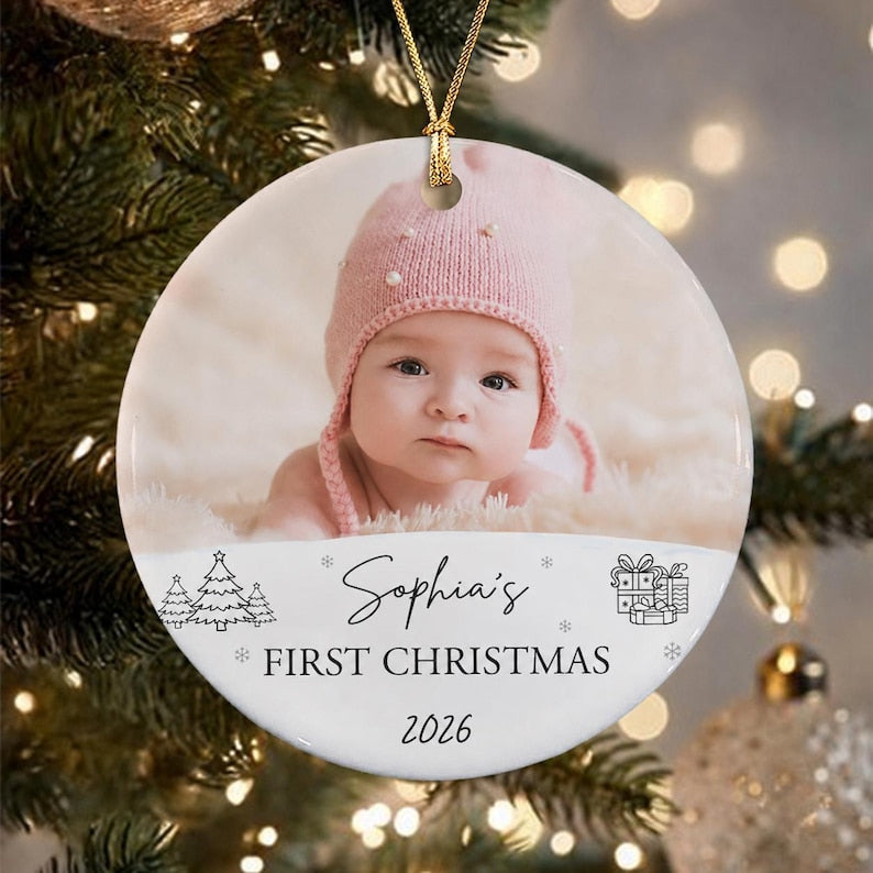 A round white ceramic ornament featuring a photograph of a baby wearing a pink knit hat. The text reads Sophia's FIRST CHRISTMAS 2026, accompanied by line art illustrations of pine trees and stacked gift boxes with bows.