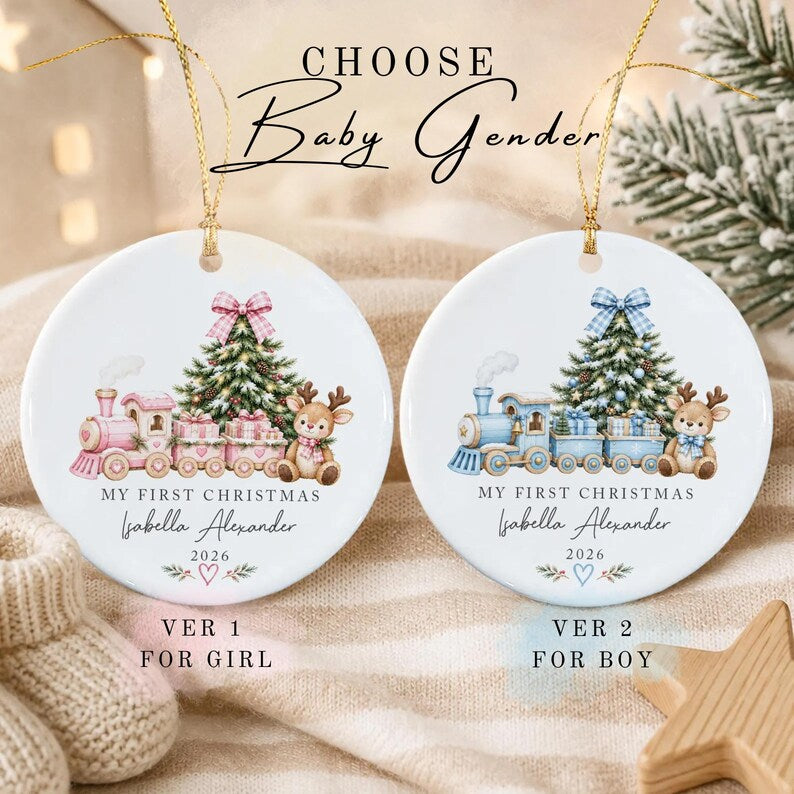 Two round white ceramic ornaments with gold hanging strings. One features a pink train, Christmas tree, and reindeer with the text 'MY FIRST CHRISTMAS Isabella Alexander 2026'.