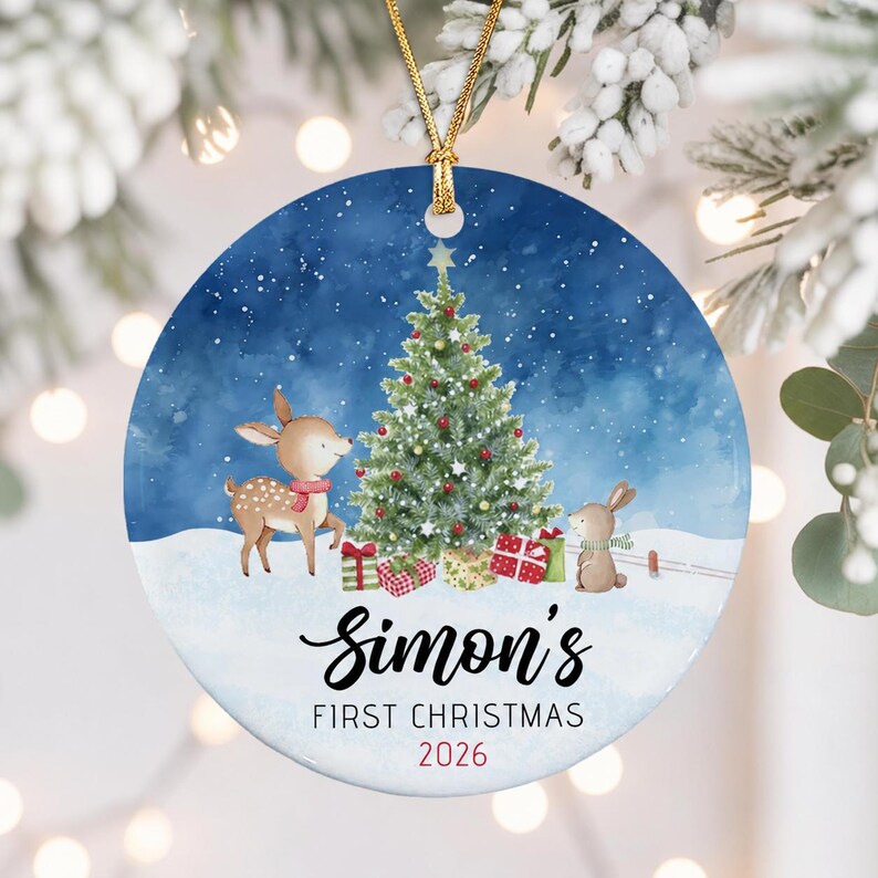 A round ceramic Christmas ornament featuring a watercolor illustration of a deer and a rabbit near a decorated tree with gifts. The text reads Simon's FIRST CHRISTMAS 2026 in black and red lettering, suspended by a gold cord.